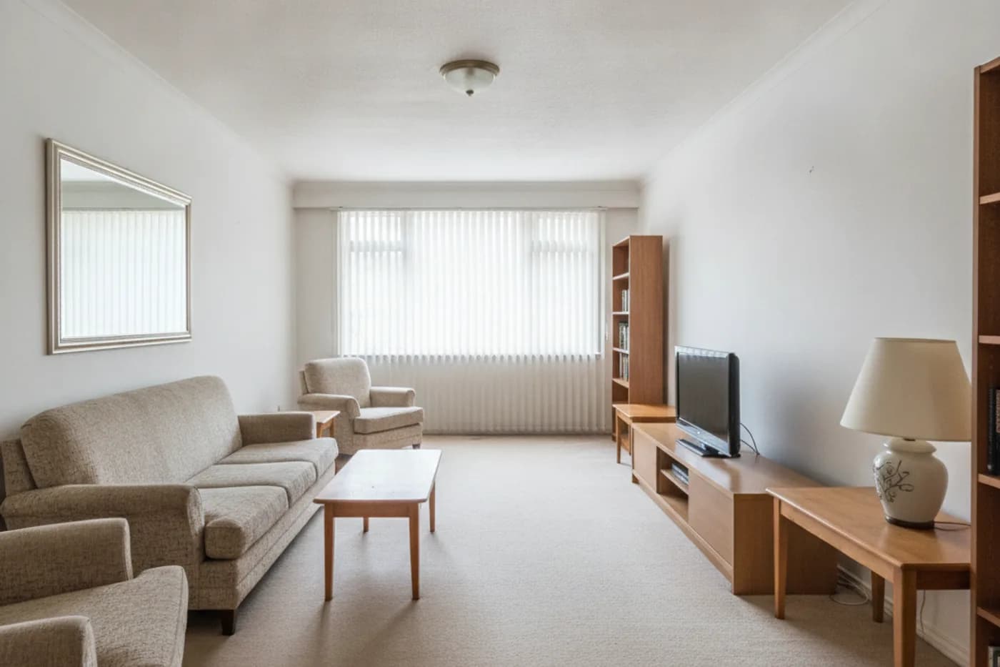 A long, narrow living room with a beige sofa and two armchairs on the left wall, a low media unit with a television on the right and a wide window at the far end